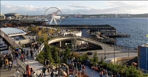 A bustling waterfront scene where people of all ages walk along sloped walkways, ramps and bridges. A ferris wheel and ferry along the shore of a large body of water in the background. In the distance are shipping cranes and cargo ships. 